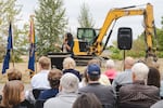 A woman speaks to a crowd at a lectern in front of an excavator.