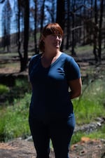 a woman with red hair and a blue t-shirt stands for a portrait in front of burnt tree skeletons and new grasses