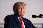 Former President Donald Trump wears a suit and red tie while standing at a podium with a microphone.