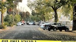 Police tape blocks a tree-lined street where cars are parked. A police car is visible in the distance.