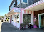 A pink and white striped building with a sign that says "Bruce's Candy Kitchen." A gumball machine stands next to the open front door of the shop, the sky is blue above without any clouds.