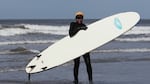 Woman walking out of ocean onto beach holding a large surfboard and smiling.
