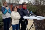 Four people standing at a podium that has media mics on it. One person in the back is keeping a kitten warm in her jacket. Grahek is speaking at the podium.