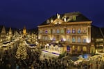 The town of Gegenbach, Germany, turns its town hall into a giant advent calendar each December.  This photo shows the front of the neo-classical town hall at an angle, with each of the 24 window shades drawn up revealing a colorful image. It is evening. There is also a Christmas tree bedecked with lights and a Christmas market in the background.