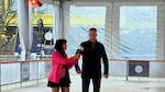 Mayor Ted Wheeler wears a long black coat and bright orange skates while being interviewed by Lillian Karabaic, who is shorter and wearing a pink blazer and pink skates.