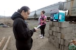 Campos looks at her phone while Castillo puts her hands on hips as they weight for a freight train to pass. A cardboard box of harm reduction supplies sits on a pile of cinderblocks.