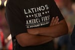 A man wearing a "Latinos for America" T-shirt stands with his arms crossed. 