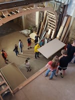 A bird's eye view of student's helping move dry wall in a team of three. Others are standing around as bystanders.