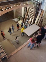 A bird's eye view of student's helping move dry wall in a team of three. Others are standing around as bystanders.