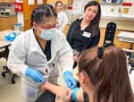 Phlebotomy student Laura Cahuich (left) preps her classmate before drawing her blood. CCC phlebotomy professor Emily Zuniga (background) supervises the students.