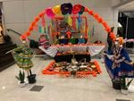An altar with pictures and candles sits in a common area of ​​the high school.  In the foreground is an ornate arch with skeletons in traditional Mexican dresses at each end.