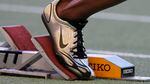 A person's foot is shown in close-up on the starting block of a race track. A Nike swoosh logo is visible on their shoe