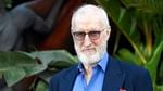 Actor James Cromwell, dressed in a blue suit jacket and shirt with a red pocket square, stands in front of a plant with large leaves. He is wearing sunglasses.