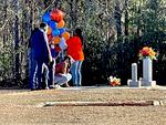 People holding colorful balloons stand together around a grave site, as one person kneels down and touches the ground.