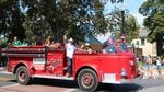 A vintage 1950s fire engine is driving down the street in a parade. Standing up front in the fire engine is Pastor Luis Molina, a man wearing a white cowboy hat, a white button up shirt, jeans and cowboy boots. He's smiling and waving toward the camera.
