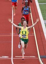 A runner in a University of Oregon yellow and green jersey raises his hands and smiles as he crosses the finish line of a track, with other runners close behind.