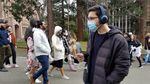 A man wearing a surgical mask and headphones walks through the University of Washington campus while holding a smartphone. People walk behind him.
